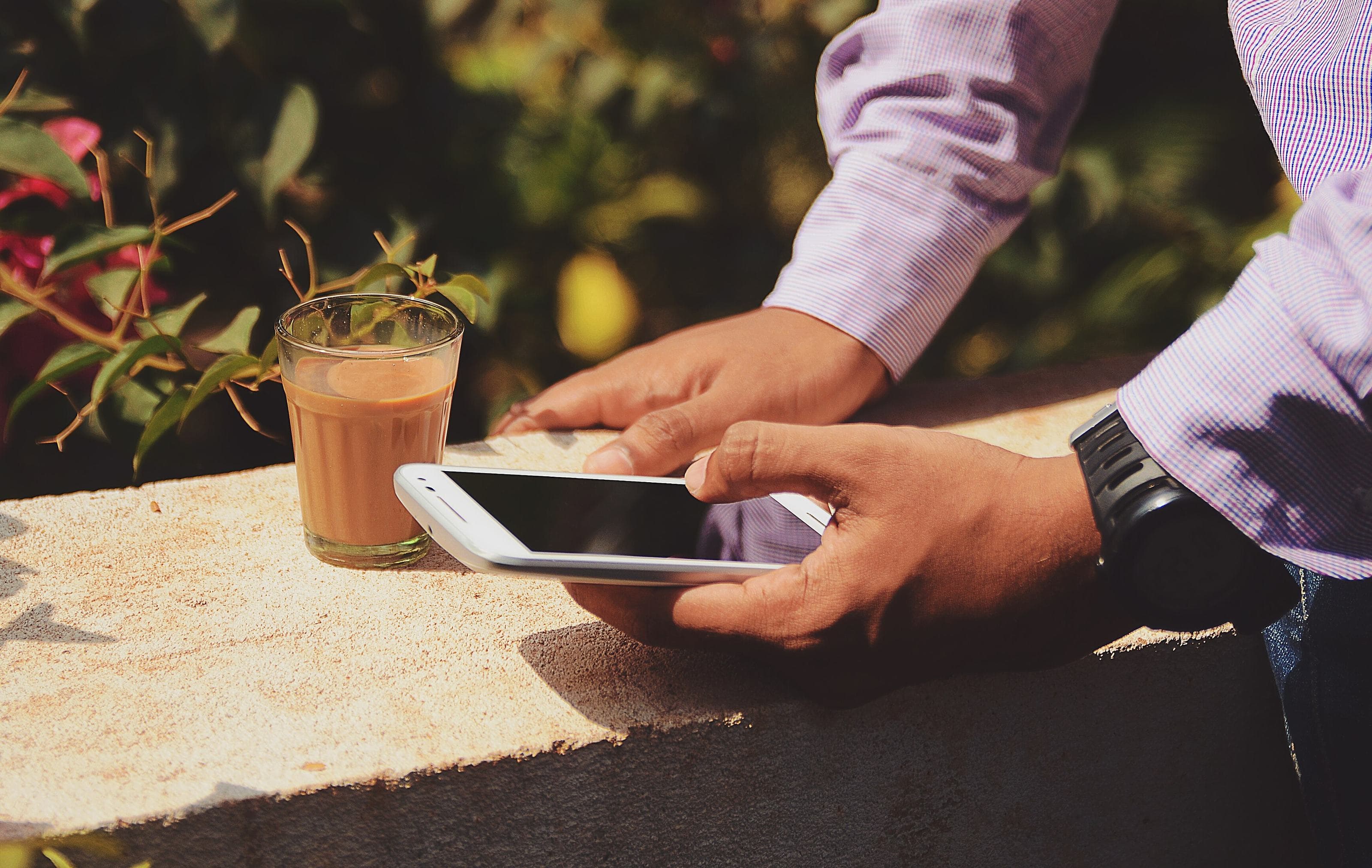 Una mano tiene il telefono di piatto sopra un muretto al sole, accanto a un bicchiere